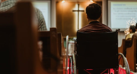 Man in a wheelchair sitting in a church pew, seen from behind, with a cross on the wall ahead