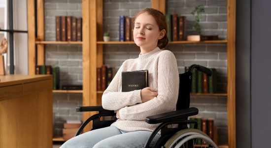 Young woman in a wheelchair with her eyes closed, holding a Holy Bible to her chest in a library