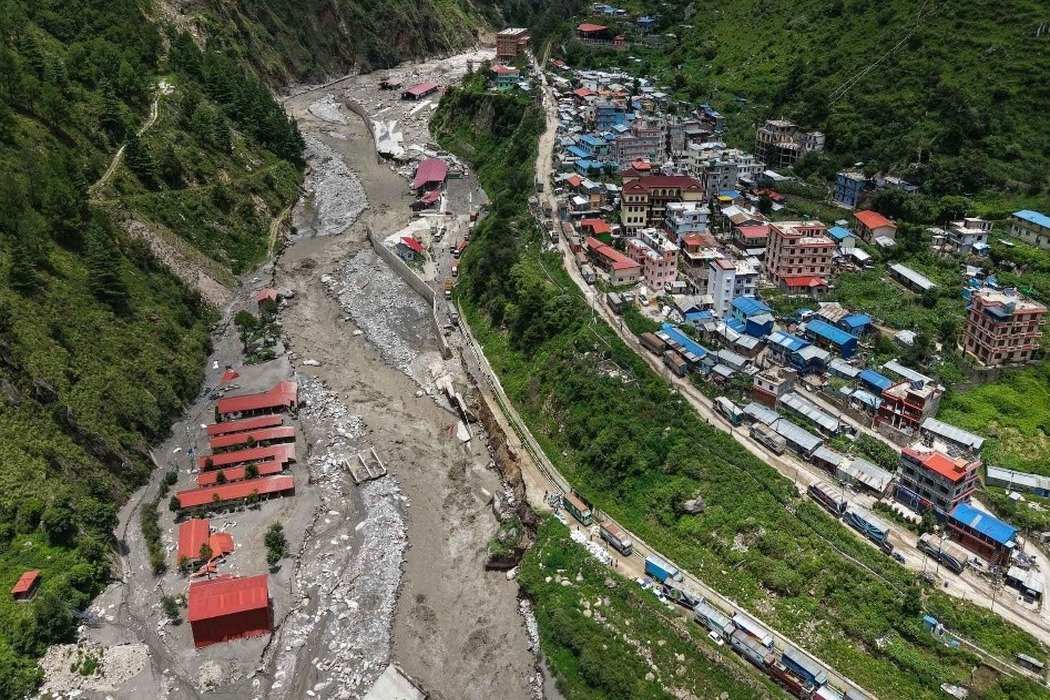An aerial view of a Nepali town beside a flood-scoured river, with vehicles queued along the damaged road.